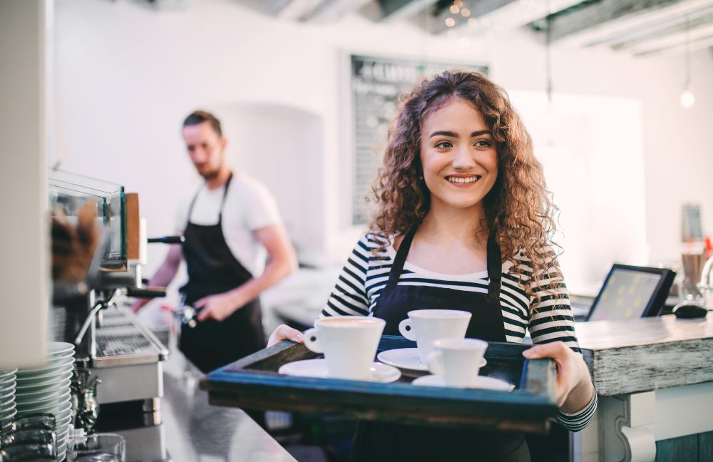 300 jobs in 100 days - image of a young woman with curly hair holding a tray of coffee cups in a cafe with barista blurred in the background