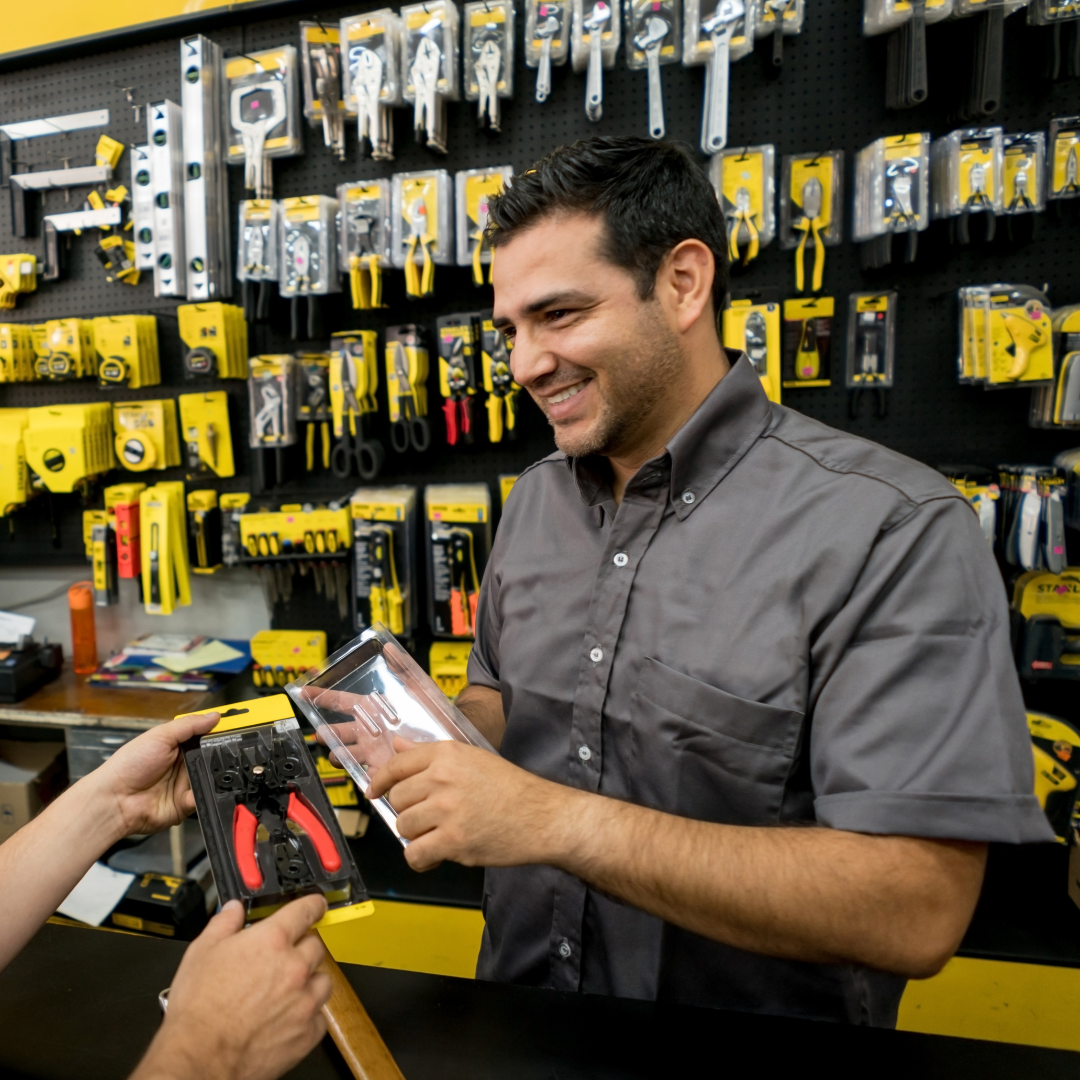 Picture of a storeman smiling at a desk, behind him is rows of tools.